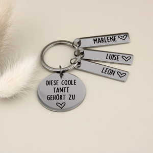 Silberner Schlüsselanhänger mit Gravur 'Diese coole Tante gehört zu' und drei rechteckigen Anhängern mit den Namen Marlene, Luise und Leon neben Trockenblumen.