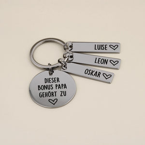 Edelstahl-Schlüsselanhänger für den Bonus Papa mit drei Namensplättchen für Luise, Leon und Oskar auf beigefarbenem Hintergrund.