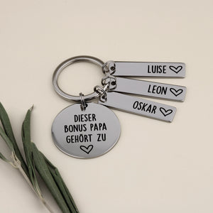 Personalisierter Schlüsselanhänger für den Bonus Papa mit Namensanhängern für Luise, Leon und Oskar neben einem frischen Olivzweig.
