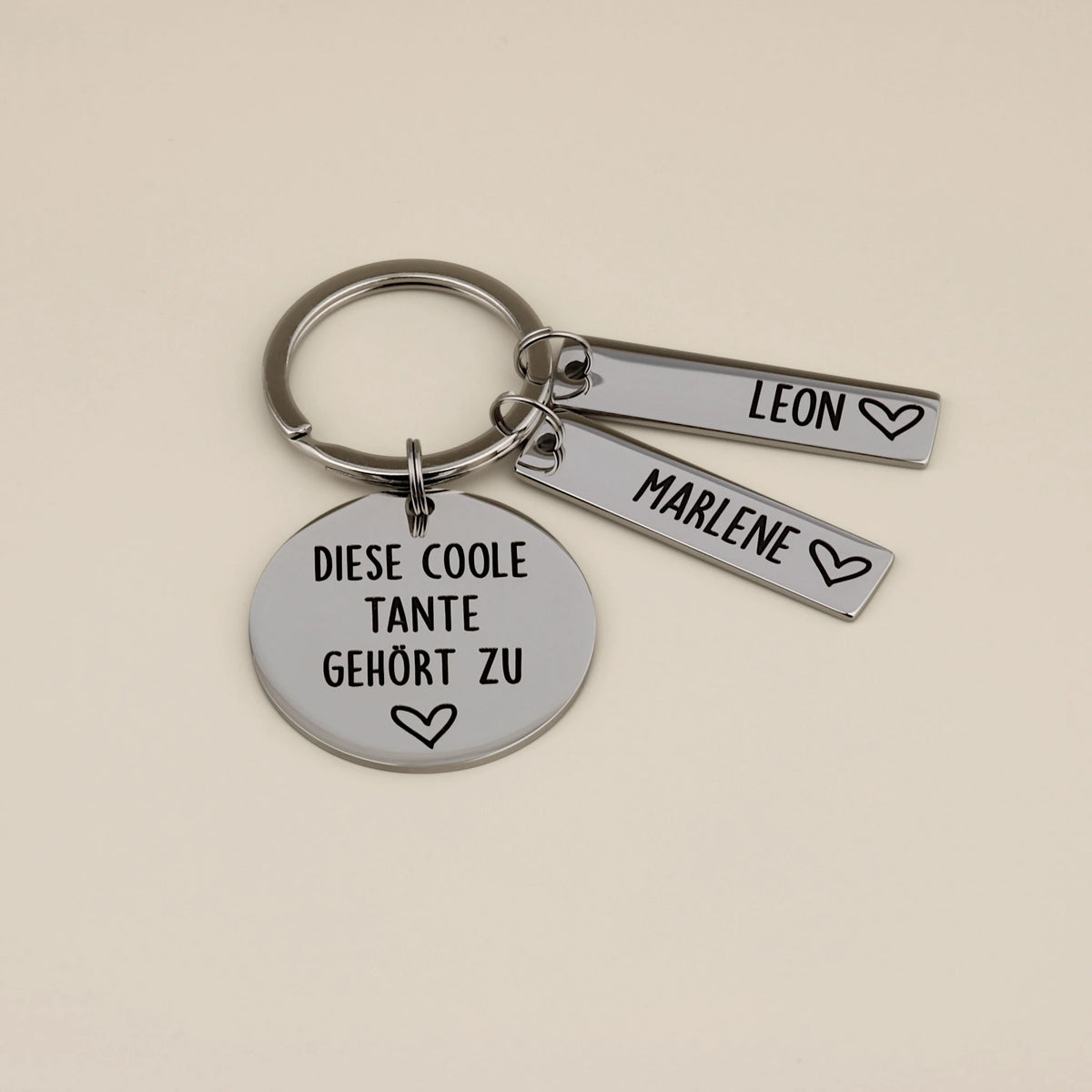 Personalisierter Schlüsselanhänger für Tante aus hochglanzpoliertem Edelstahl mit der Gravur 'Diese coole Tante gehört zu' und zwei Namensanhängern für Leon und Marlene auf beigefarbenem Hintergrund.
