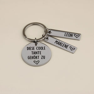 Personalisierter Schlüsselanhänger für Tante aus hochglanzpoliertem Edelstahl mit der Gravur 'Diese coole Tante gehört zu' und zwei Namensanhängern für Leon und Marlene auf beigefarbenem Hintergrund.
