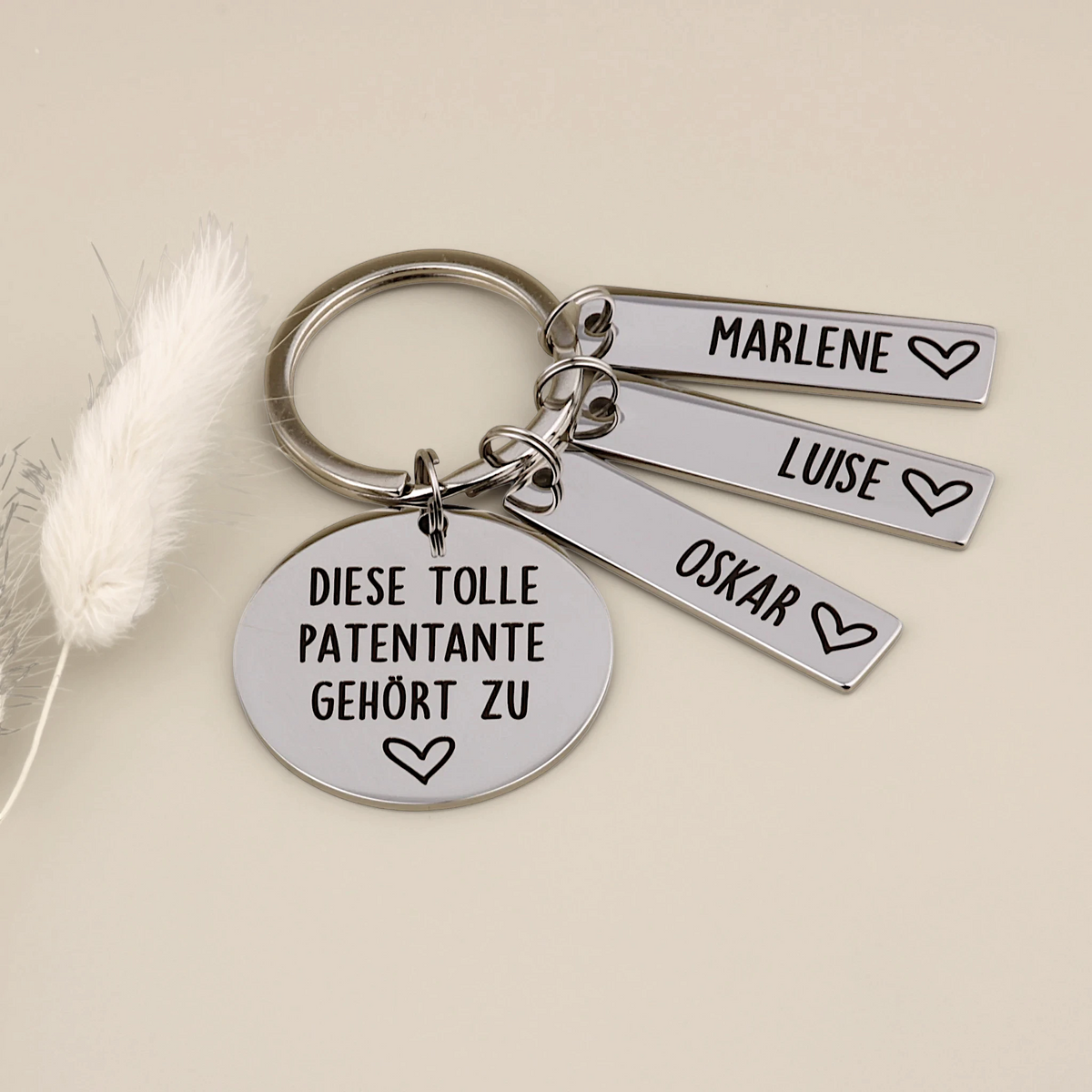 Schlüsselanhänger „Diese tolle Patentante gehört zu“ mit drei Namensanhängern neben hellen Trockenblumen.