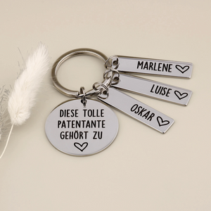 Schlüsselanhänger „Diese tolle Patentante gehört zu“ mit drei Namensanhängern neben hellen Trockenblumen.