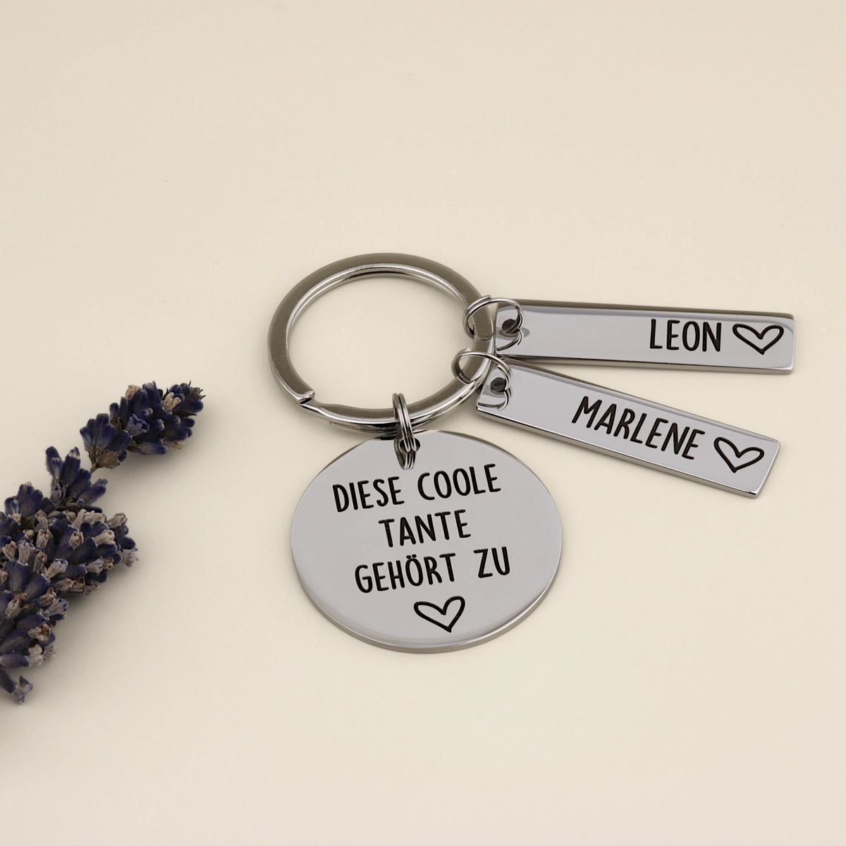 Personalisierter Schlüsselanhänger für Tante aus Edelstahl mit Gravur 'Diese coole Tante gehört zu' und zwei Namensanhängern für Leon und Marlene, dekoriert mit Lavendel.
