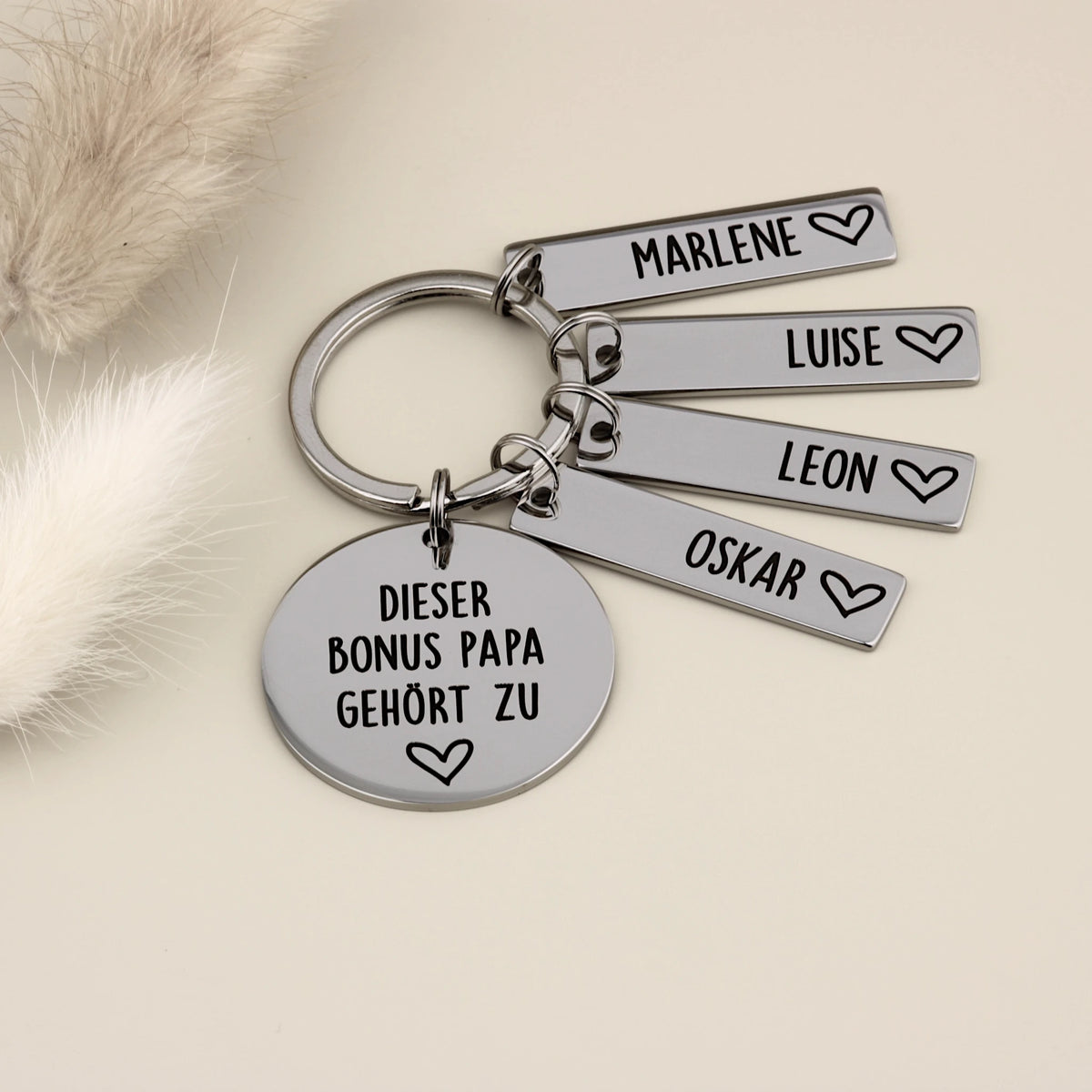 Edelstahl-Schlüsselanhänger für den Bonus Papa mit vier gravierten Namensanhängern für Marlene, Luise, Leon und Oskar, neben Trockenblumen.
