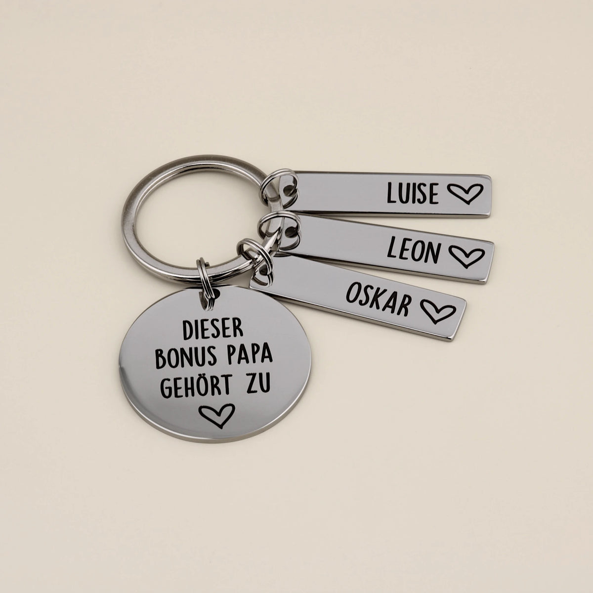 Edelstahl-Schlüsselanhänger für den Bonus Papa mit drei Namensplättchen für Luise, Leon und Oskar auf beigefarbenem Hintergrund.
