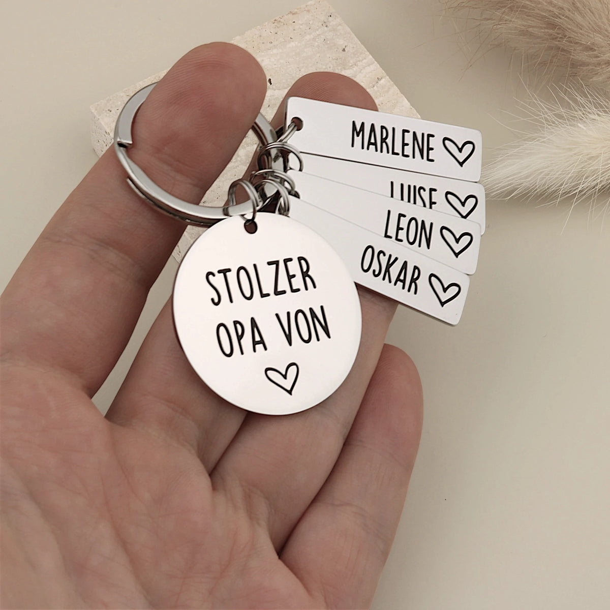 Person hält einen personalisierten Stolzer Opa-Schlüsselanhänger mit vier Namensanhängern als Größenvergleich.
