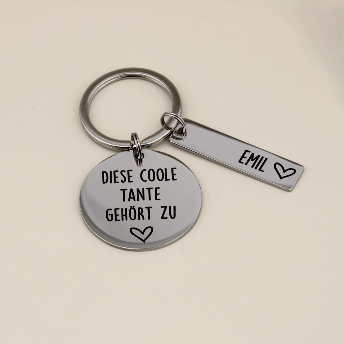 Nahaufnahme des gravierten Schlüsselanhängers 'Diese coole Tante gehört zu' mit einem Namensanhänger für Emil.
